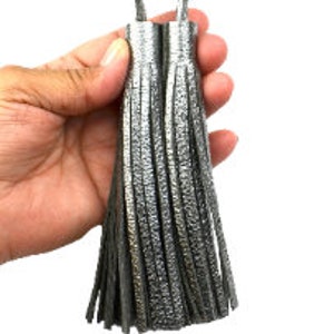 May include: Two metallic silver tassels featuring a textured finish and long, thin fringe strands. Each tassel is topped with a cylindrical cap and includes a matching silver loop at the top for attachment, showcasing a reflective surface.