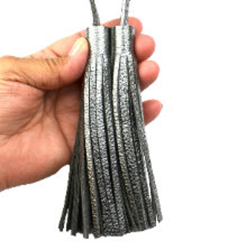 May include: Two metallic silver tassels featuring a textured finish and long, thin fringe strands. Each tassel is topped with a cylindrical cap and includes a matching silver loop at the top for attachment, showcasing a reflective surface.