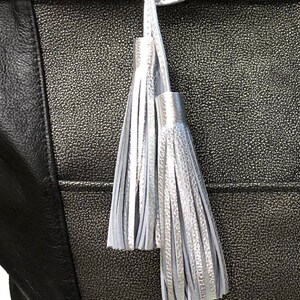 May include: A black tote bag featuring a textured metallic silver center panel and a smooth black side panel. Two silver tassels hang from the top handle, attached by a silver loop. The bag has a distinct pebbled finish on the metallic section.