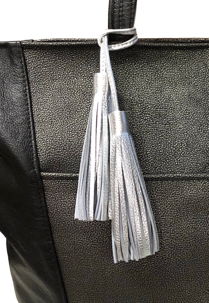 May include: A black tote bag featuring a textured metallic silver center panel and a smooth black side panel. Two silver tassels hang from the top handle, attached by a silver loop. The bag has a distinct pebbled finish on the metallic section.