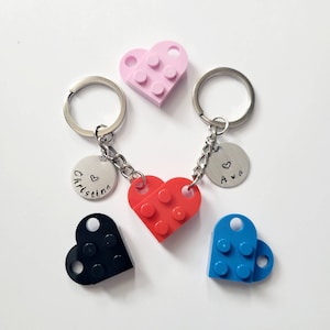 May include: Keyrings featuring heart-shaped Lego brick designs in red, pink, black, and blue. Each keyring has a silver ring and a small, round charm with a name and a heart symbol. A unique gift idea.