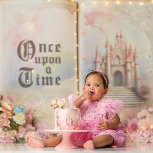 May include: A baby girl in a pink dress sits in front of a backdrop depicting a fairytale castle and the words "Once upon a time". She is holding a pink and white cake with a castle design. The backdrop is decorated with pink flowers and lights.