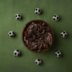 May include: A wooden bowl filled with brown and white shredded material, surrounded by eight black and white footballs on a green background.