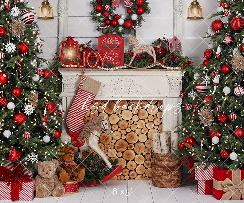 May include: A festive Christmas backdrop featuring two decorated trees with red and white ornaments, and a fireplace with a stocking, rocking horse, and the word "JOY". The backdrop includes wrapped gifts and a "Letters to Santa" mailbox.