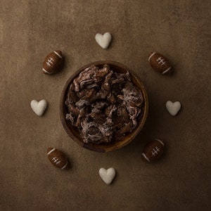 May include: A brown wooden bowl filled with brown and white textured material. The bowl is surrounded by white felt hearts and brown and white footballs.