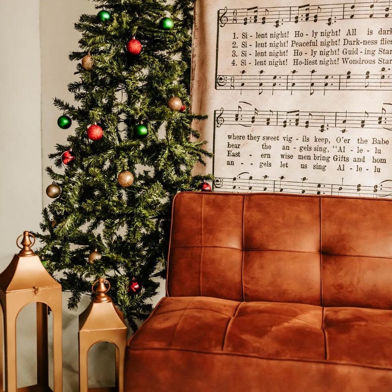 May include: A decorated Christmas tree with red, green, and gold ornaments stands next to a brown leather couch. Two gold lanterns are in the foreground. Sheet music with the lyrics "Silent Night" is visible in the background.