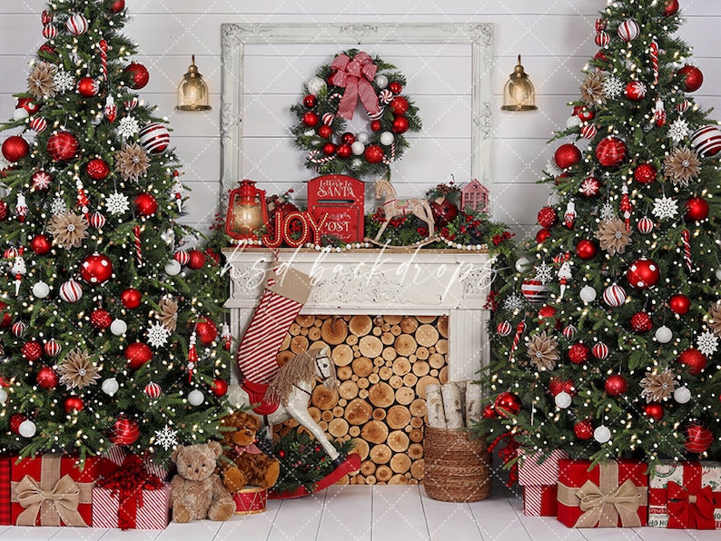 May include: A Christmas-themed backdrop featuring two decorated trees with red and white baubles and wrapped presents. A fireplace mantel displays a wreath, stocking, and rocking horse. The word "JOY" is spelled out in decorative letters. A "Letters to Santa" mailbox is also present.