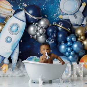 May include: A baby boy sits in a white bathtub with a blue and silver balloon arch behind him. The baby is holding a white washcloth in his mouth and looking at the camera. The balloons are in the shape of planets and stars. The background is a dark blue with white stars. The word "ONE" is spelled out in white lights in front of the bathtub.