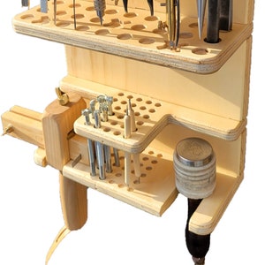 May include: A wooden tool organizer with multiple shelves and compartments for storing leatherworking tools. The organizer has a variety of tools, including awls, punches, and a hammer.