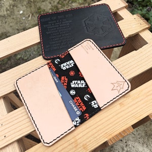 May include: A light brown leather wallet with a black and red Star Wars themed fabric insert. The wallet has a stitched edge and a small star design on the front. The wallet is sitting on a wooden surface.