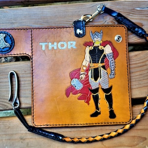 Puede incluir: Una cartera de cuero marrón con una correa trenzada negra. La cartera tiene un diseño de personaje de Thor y la palabra "THOR" en relieve en la parte delantera. La cartera también tiene un símbolo de martillo de metal en la parte delantera.