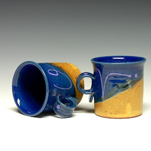 May include: Two blue ceramic mugs with a yellow glaze on the bottom. The mugs have a purple swirl design and are handmade.