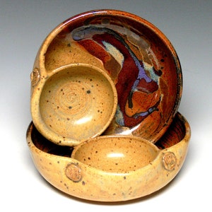 May include: Three ceramic bowls nested together. The top bowl is a deep brown with a swirl of purple and white glaze. The middle bowl is a light brown with a speckled glaze. The bottom bowl is a light brown with a speckled glaze and three raised knobs.