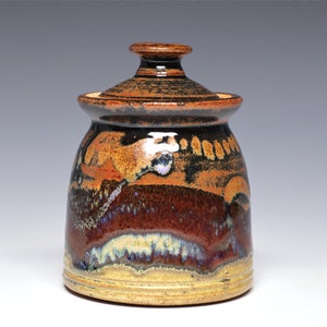 May include: A ceramic jar with a lid, featuring a brown, black, and white glaze. The jar has a rounded body and a slightly flared rim.