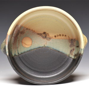 May include: Handmade ceramic bowl with a unique color gradient. The bowl features a dark gray base that transitions to a light green, brown, and beige interior. Two small handles are on the sides. The bowl has a rustic, artistic aesthetic.