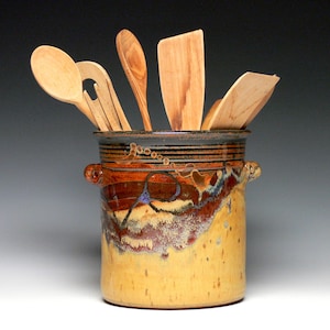 May include: A ceramic utensil holder with a yellow, brown, and black design, holding several wooden spoons and spatulas. The holder has two small handles and a decorative pattern around the middle. The wooden utensils are various shapes and sizes.