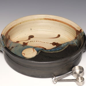 May include: A handmade ceramic baking dish with a black exterior and a cream-colored interior with blue and brown abstract designs. Two stainless steel measuring spoons are next to the dish.