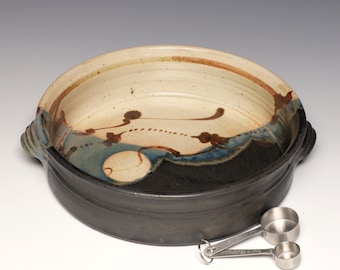 May include: A handmade ceramic baking dish with a black exterior and a cream-colored interior with blue and brown abstract designs. Two stainless steel measuring spoons are next to the dish.