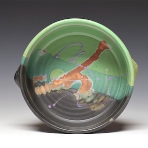 May include: A handmade ceramic bowl with a green interior and a black exterior. The bowl features abstract brown and purple painted designs. The bowl has two handles and is suitable for serving food.