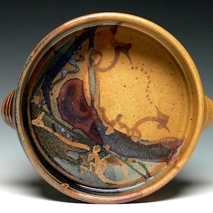 May include: A handmade ceramic bowl with a round shape and two handles. The bowl features a speckled tan interior with abstract designs in shades of brown, orange, purple, and black. The rim and handles are a darker brown.