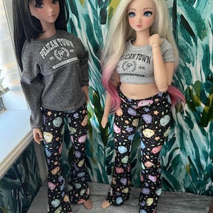 May include: Two dolls dressed in matching outfits. One doll wears a grey long-sleeved top, the other a grey crop top, both with "Pelican Town" text. Both dolls are wearing black flared trousers with colourful fruit and star patterns.