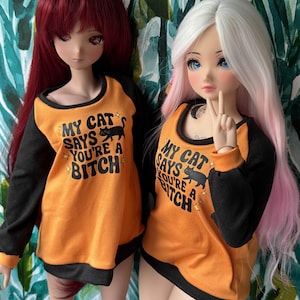 以下が含まれることがあります： オレンジと黒の長袖シャツを着た2体のアニメ風人形。シャツには「MY CAT SAYS YOU'RE A BITCH」の文字と黒猫のグラフィックが入っています。1体は赤毛、もう1体はピンクのハイライトが入った白髪です。