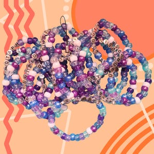 May include: A collection of colorful beaded bracelets in shades of purple, blue, and white. Some beads have silver letter beads spelling out words. The bracelets are strung together and have a black string attached.