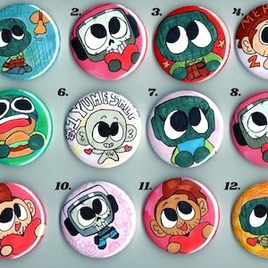 May include: A collection of twelve colorful, round pinback buttons. Each button features a unique cartoon character design, with various expressions and themes. Some buttons include text like "MEAN 2 ME" and "YUME STILL".