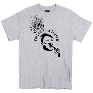 May include: A light gray t-shirt featuring a black graphic of a person holding a gun to their mouth, with the text "FOLLOW YOUR LEADER" in a circular design. Above the graphic is a black splatter design.