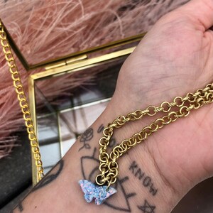 Può includere: Una collana a catena dorata con un ciondolo a forma di farfalla blu scintillante. La collana è sul polso di una persona con un tatuaggio che dice "know".