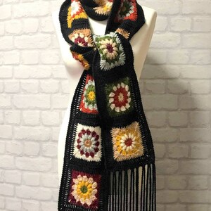 Pode incluir: Uma echarpe de croch&ecirc; preta com motivos florais coloridos e franjas. A echarpe &eacute; feita de fio e tem um design de quadrado da av&oacute;.