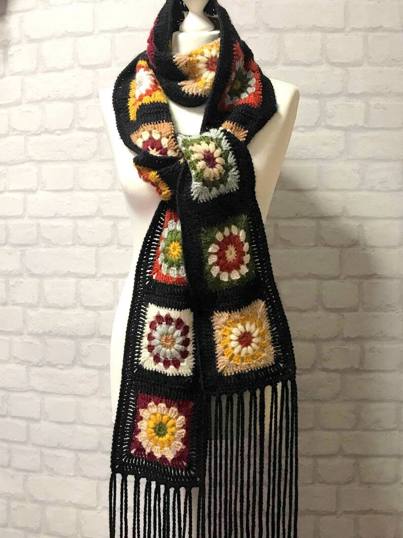 Pode incluir: Uma echarpe de croch&ecirc; preta com motivos florais coloridos e franjas. A echarpe &eacute; feita de fio e tem um design de quadrado da av&oacute;.