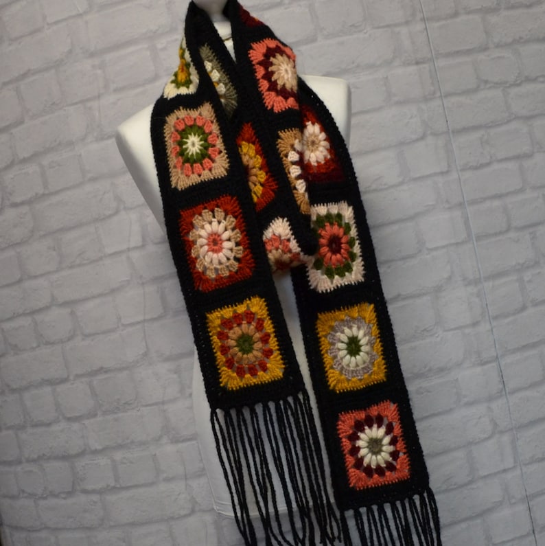 Pode incluir: Uma echarpe de croch&ecirc; preta com quadrados de av&oacute; coloridos e franjas pretas. A echarpe &eacute; feita com uma variedade de cores, incluindo vermelho, laranja, amarelo, verde e marrom.