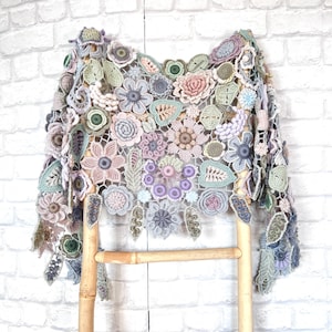 May include: A crocheted shawl with a floral design in shades of pink, purple, green, and gray. The shawl is draped over a wooden ladder.