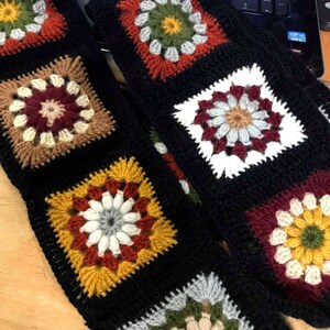 Pode incluir: Uma echarpe de croch&ecirc; preta com quadrados de av&oacute; coloridos. Os quadrados apresentam um desenho de flores em tons de vermelho, branco, amarelo, verde e marrom.