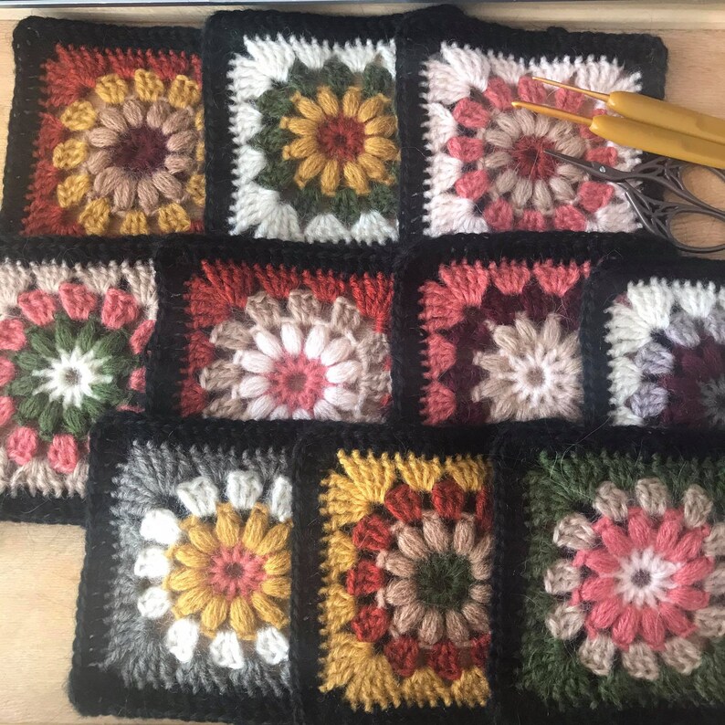 Pode incluir: Nove quadrados de croch&ecirc; com flores de diferentes cores no centro. Os quadrados s&atilde;o emoldurados com fio preto e est&atilde;o dispostos em tr&ecirc;s filas.