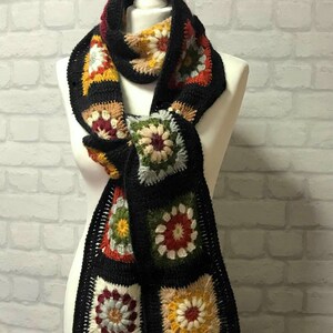 Pode incluir: Uma echarpe de croch&ecirc; preta com quadrados de av&oacute; coloridos que apresentam um design floral. A echarpe tem franjas pretas nas pontas.