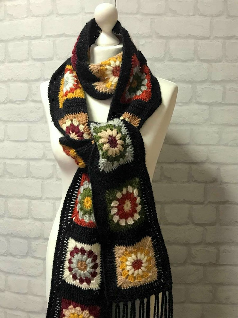 Pode incluir: Uma echarpe de croch&ecirc; preta com quadrados de av&oacute; coloridos que apresentam um design floral. A echarpe tem franjas pretas nas pontas.