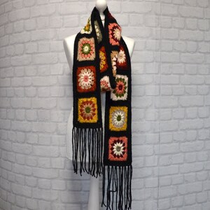 Pode incluir: Uma echarpe de croch&ecirc; preta com quadrados florais coloridos e franjas.