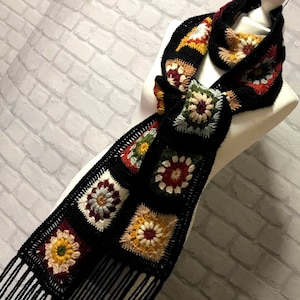 Pode incluir: Uma echarpe de croch&ecirc; preta com motivos coloridos de quadrado da av&oacute; e franjas pretas. A echarpe &eacute; exibida em um manequim branco.