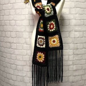 Pode incluir: Uma echarpe de croch&ecirc; preta com quadrados de av&oacute; coloridos e franjas pretas. A echarpe &eacute; exibida em um manequim branco contra uma parede de tijolo branco.