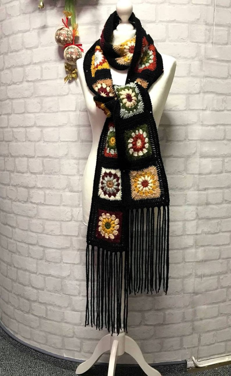 Pode incluir: Uma echarpe de croch&ecirc; preta com quadrados de av&oacute; coloridos e franjas pretas. A echarpe &eacute; exibida em um manequim branco contra uma parede de tijolo branco.