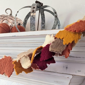 May include: A decorative garland of felt leaves in autumnal colours: orange, yellow, cream, and burgundy. The garland is draped over a white mantelpiece, with pumpkins and a metal crown-shaped object on the shelf above.