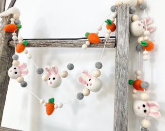 May include: Easter garland featuring white felt bunny heads with pink ears and cheeks, orange felt carrots with green tops, and wooden beads. The garland is strung on white string and is displayed on a wooden ladder.