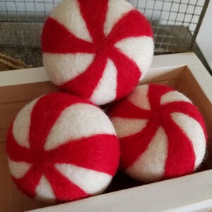 K&ouml;nnte beinhalten: Drei rote und wei&szlig;e Filzkugeln mit Pfefferminz-Muster in einer wei&szlig;en Holzkiste. Die Kugeln sind aus Wollfilz und haben eine weiche, flauschige Textur.