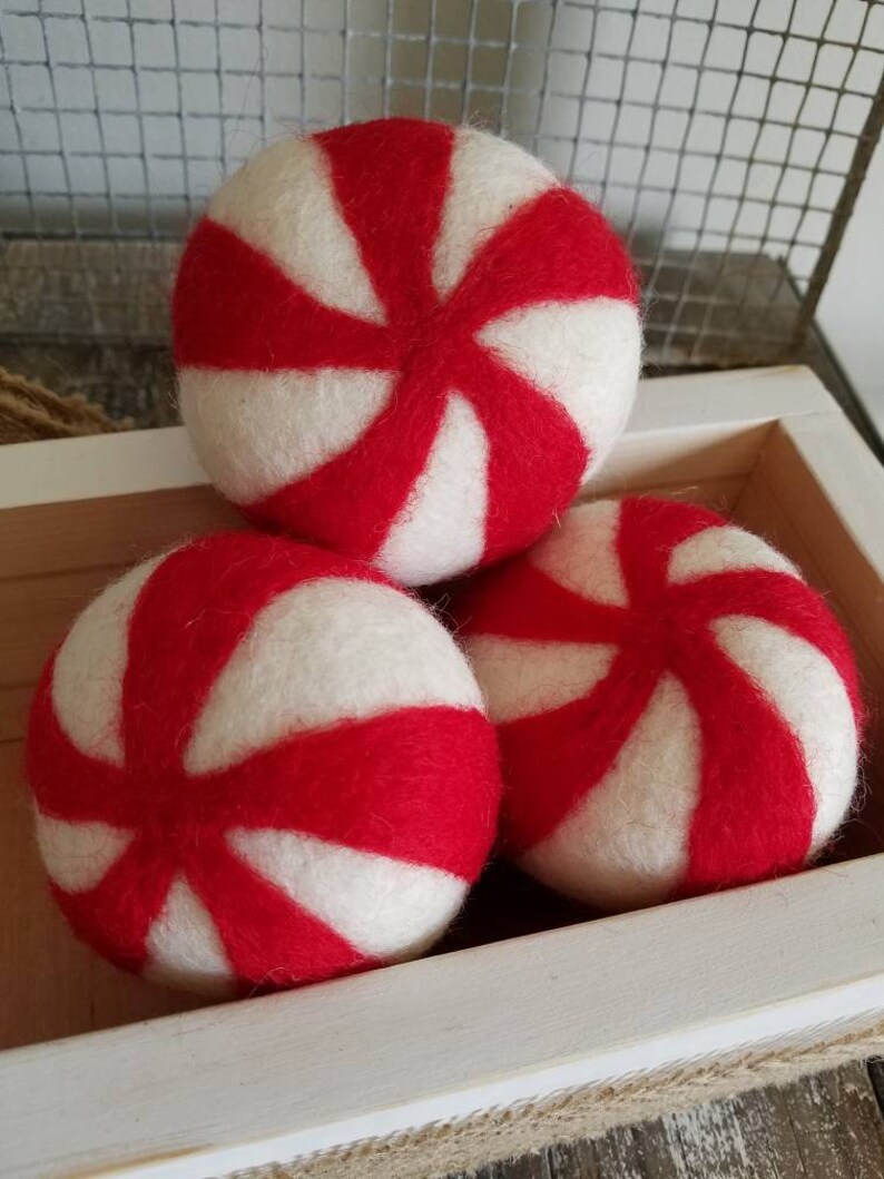 K&ouml;nnte beinhalten: Drei rote und wei&szlig;e Filzkugeln mit Pfefferminz-Muster in einer wei&szlig;en Holzkiste. Die Kugeln sind aus Wollfilz und haben eine weiche, flauschige Textur.
