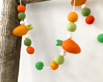 May include: A felt garland featuring orange carrots and green and orange felt balls, interspersed with wooden beads. The garland is strung on a white cord, perfect for Easter or spring decor.