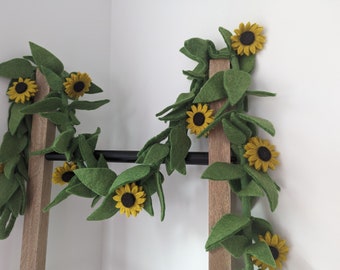 Könnte beinhalten: Eine Filzgirlande mit grünen Blättern und gelben Filzsonnenblumen. Die Girlande ist über eine Holzleiter gelegt.