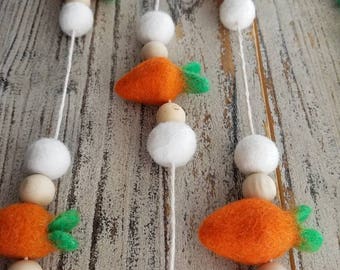 May include: A decorative garland featuring orange felt carrots with green tops, white felt balls, and wooden beads strung on white twine. The garland is designed for Easter or spring decor.