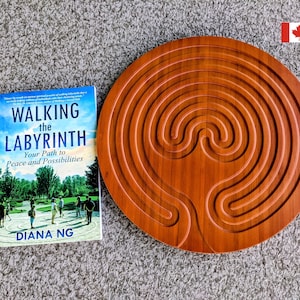 May include: A wooden labyrinth with concentric circular pathways sits next to a book titled "Walking the Labyrinth: Your Path to Peace and Possibilities" by Diana Ng. The labyrinth is a warm brown color, and the book has a blue and white cover.